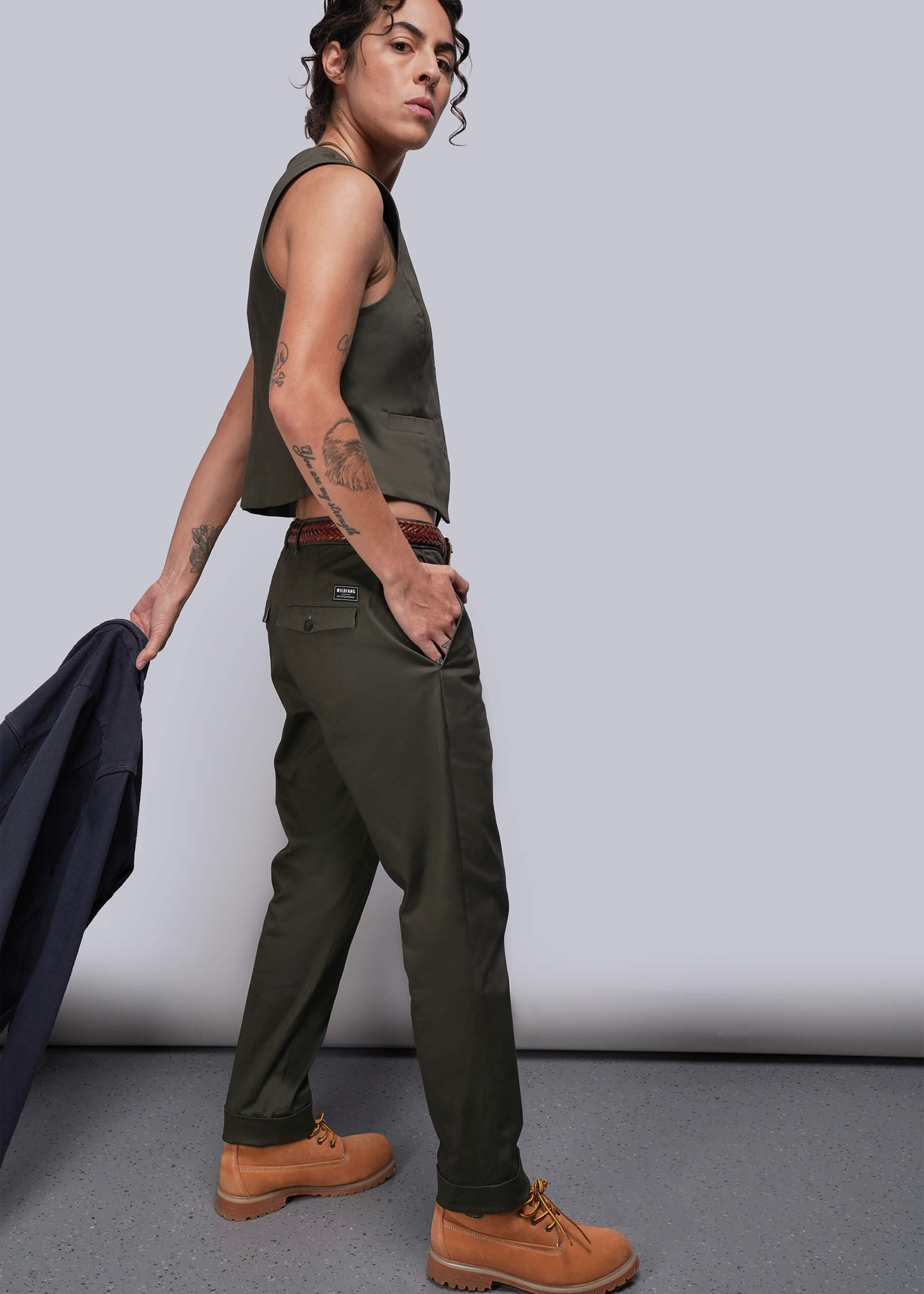 S=Person wearing a green vest and pants with brown boots, holding a jacket against a plain background.