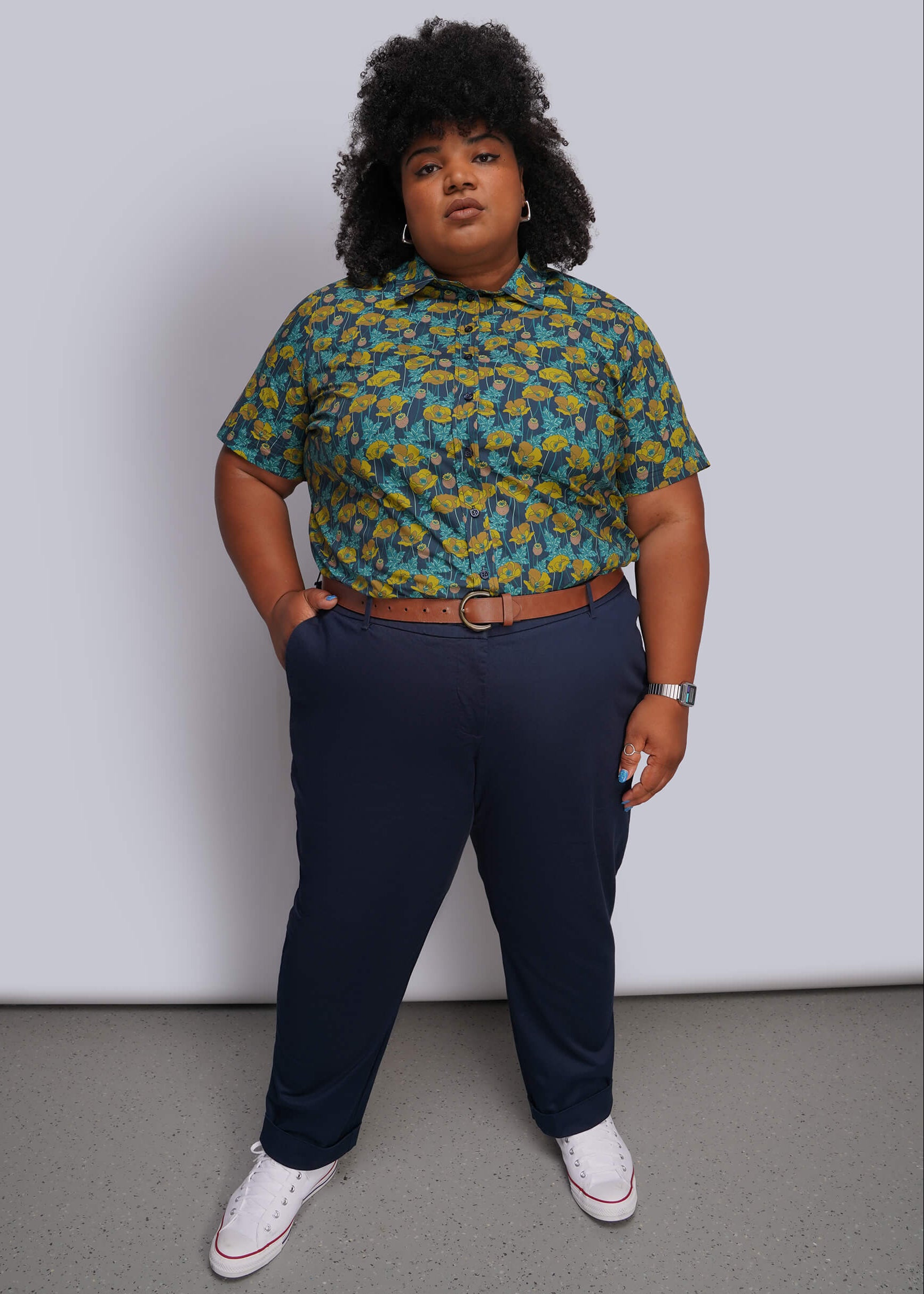 1X=A person modeling a short-sleeve button-up shirt with a green and yellow abstract pattern, paired with navy blue trousers, a brown belt, and white sneakers, standing against a light gray wall.