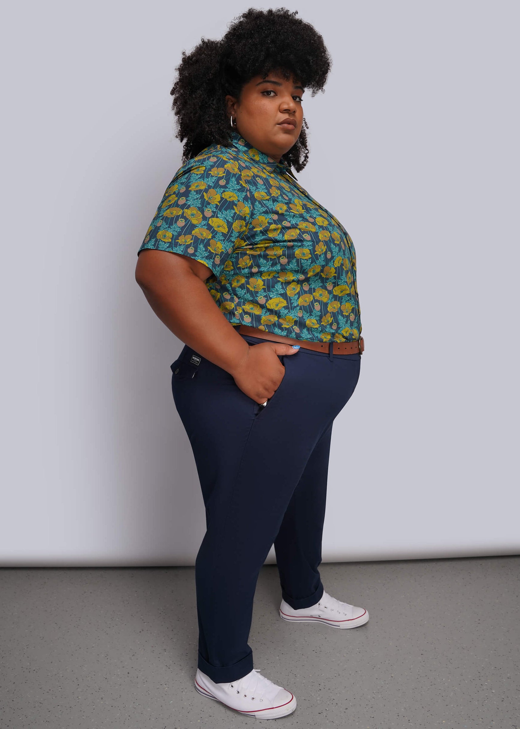 1X=A person modeling a short-sleeve button-up shirt with a green and yellow floral pattern, paired with navy blue trousers and white low-top sneakers, standing against a light gray studio background.