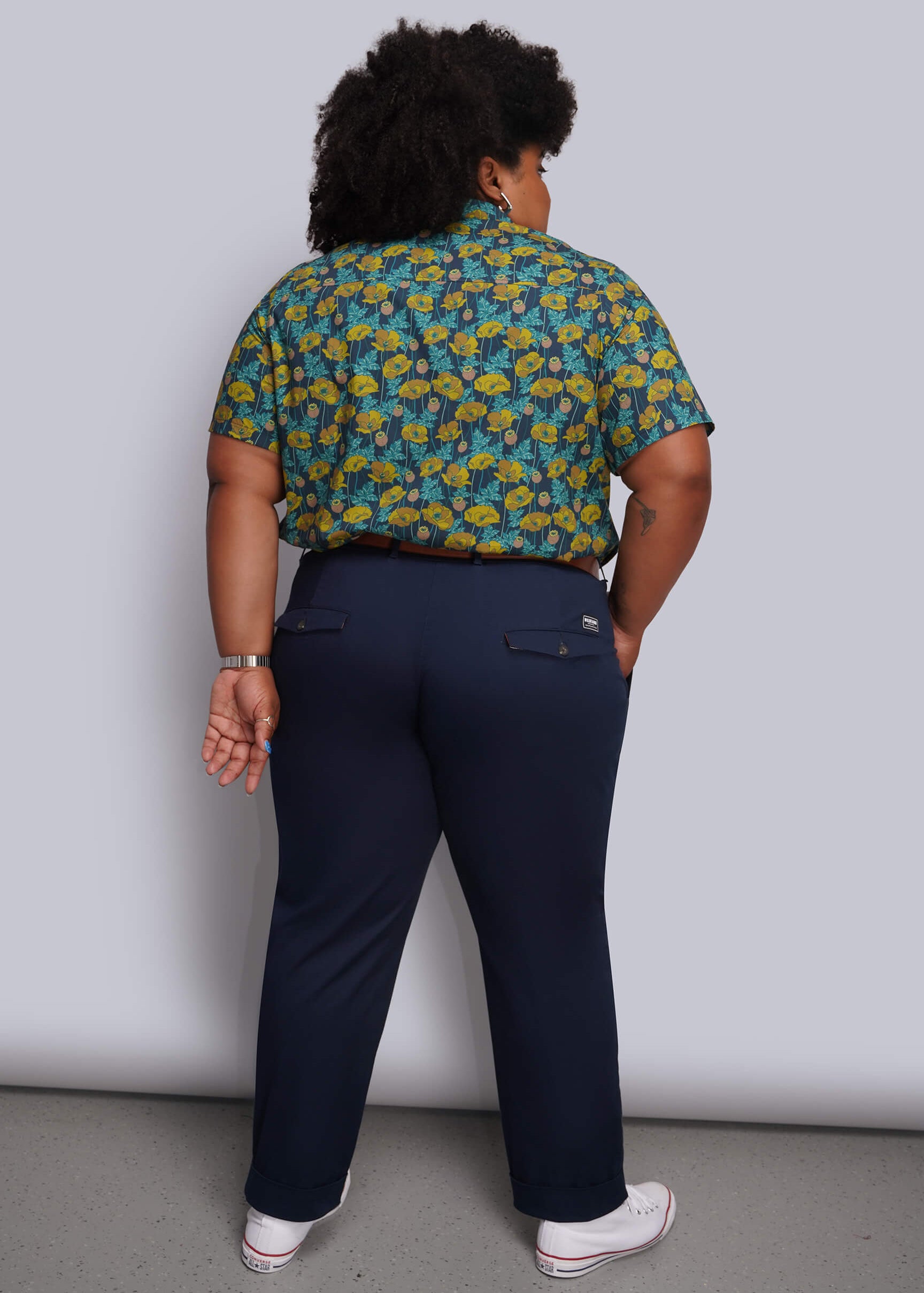 1X=A person shown from the back wearing a short-sleeve button-up shirt with a green and yellow floral pattern, navy blue trousers, and white sneakers, standing against a light gray studio wall.