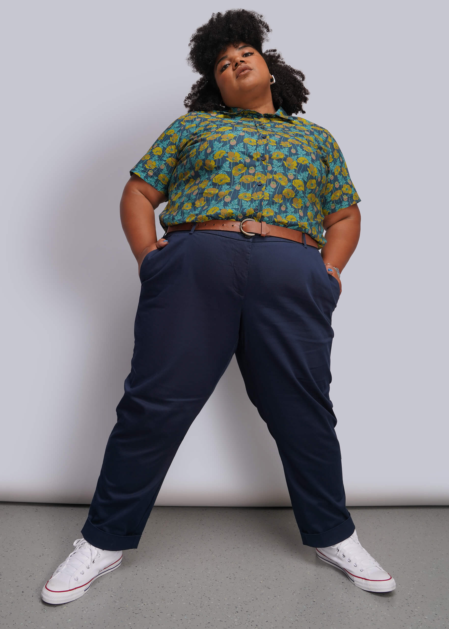 1X=A person modeling a short-sleeve button-up shirt with a green and yellow floral pattern, paired with navy blue trousers and white low-top sneakers, standing against a light gray studio background.
