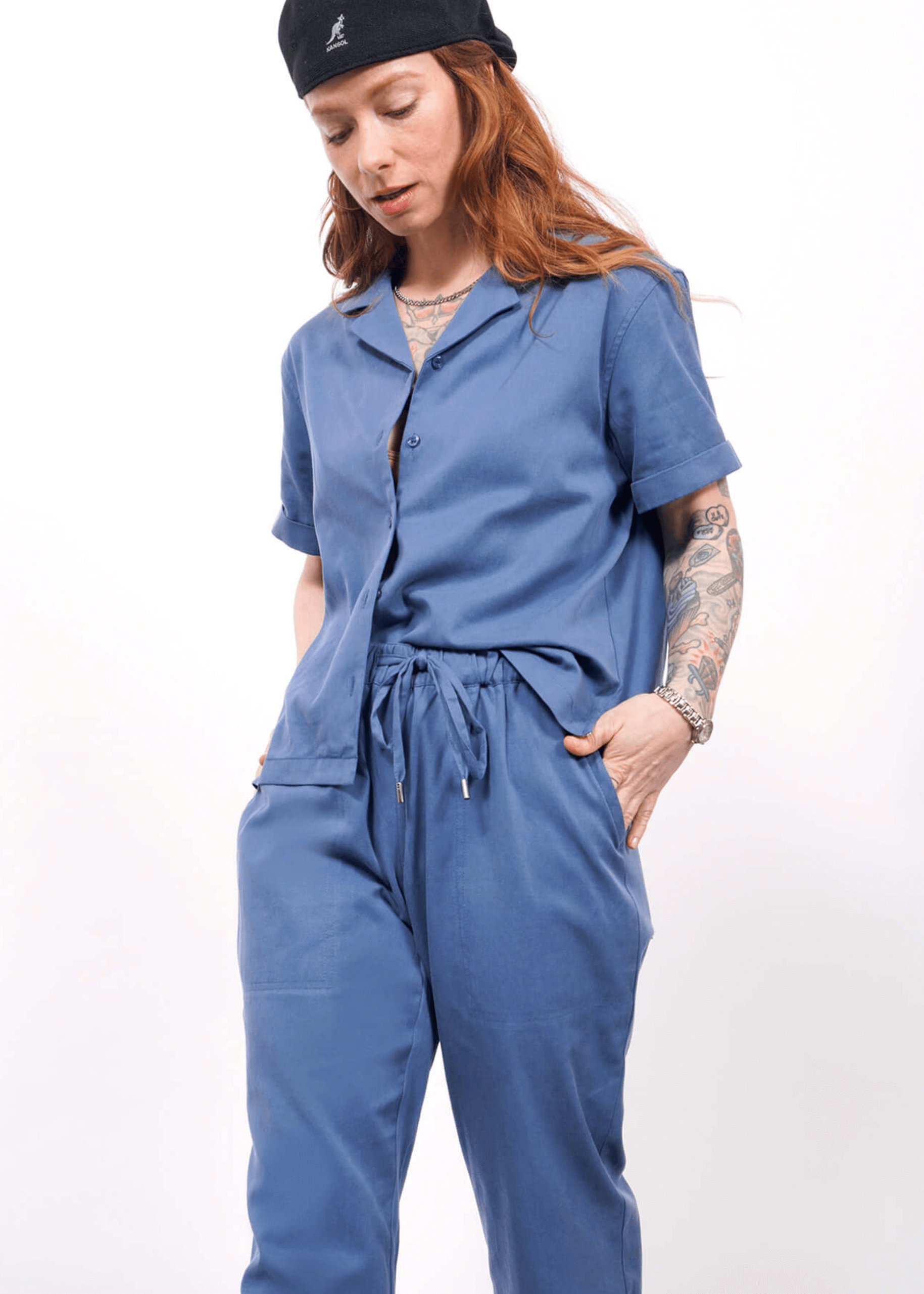 A person with long, wavy hair and visible tattoos wears a black cap and The Everyday Camp Button Up in oxford blue with matching pants, standing against a white background.