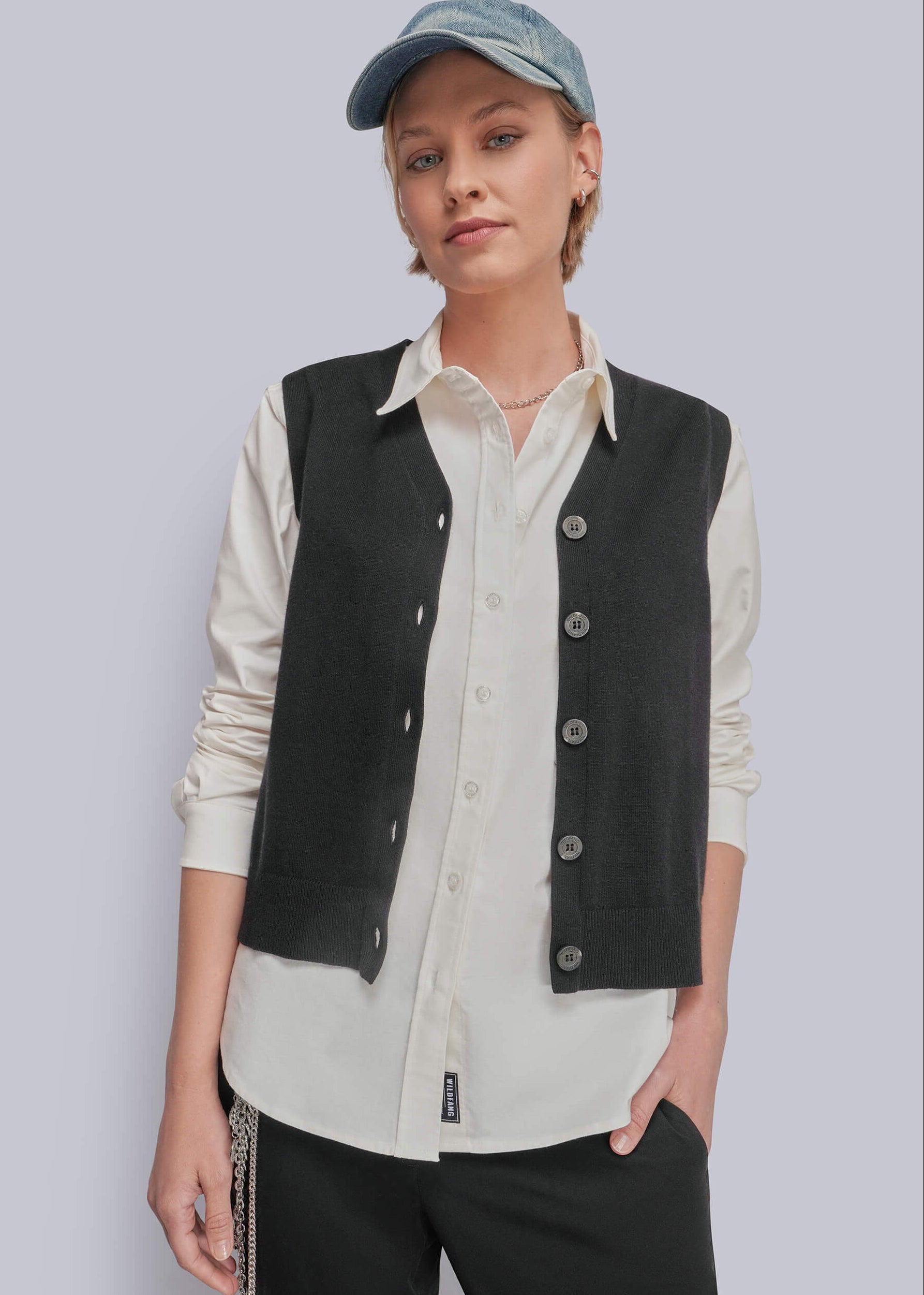 A model wearing a black button-up vest over a cream button-up shirt, paired with black pants and a blue denim cap, against a solid light gray background.