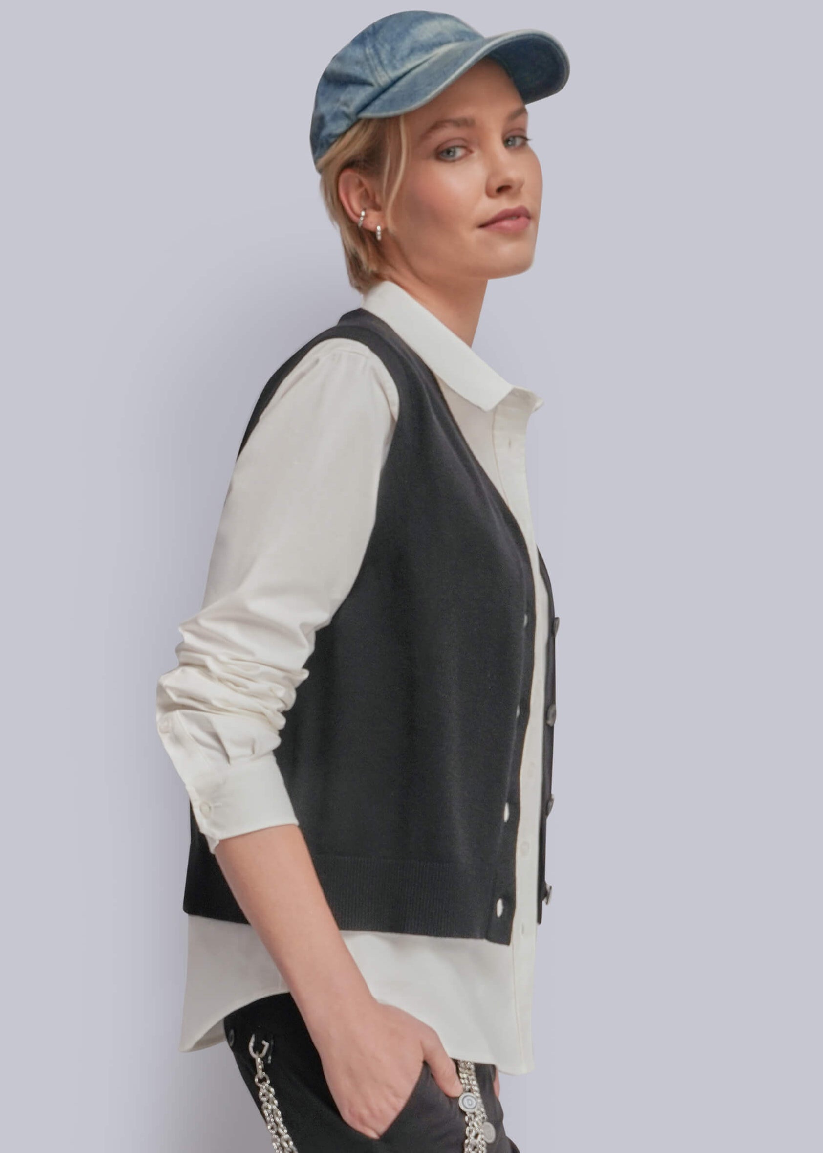 S=A woman wearing a black knit vest over a cream button-up shirt, paired with a blue denim cap and dark pants, photographed against a light gray studio background.
