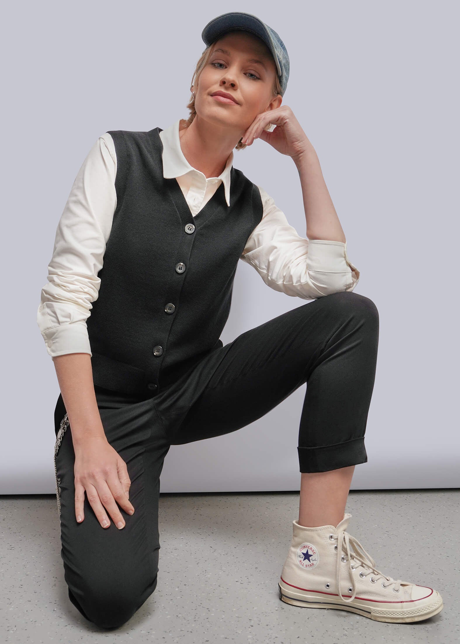 S=A model kneels on a gray floor against a light gray wall, wearing a black vest over a cream shirt, black cropped pants, and a black cap. She is wearing off-white high-top sneakers with a Converse star logo visible on the side.