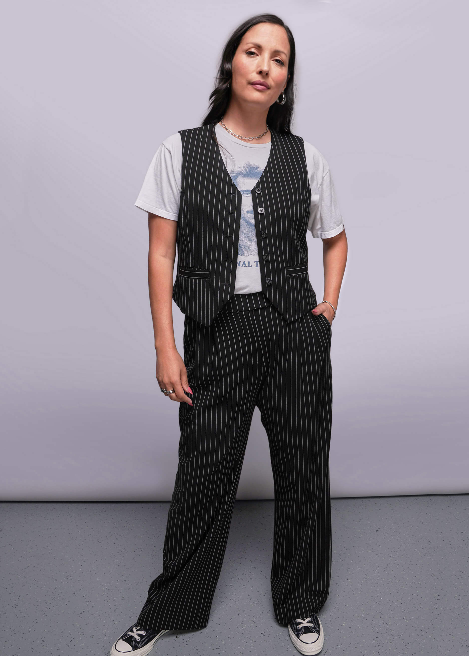 Model stands in front of gray background wearing pinstripe vest over t shirt and trouser 