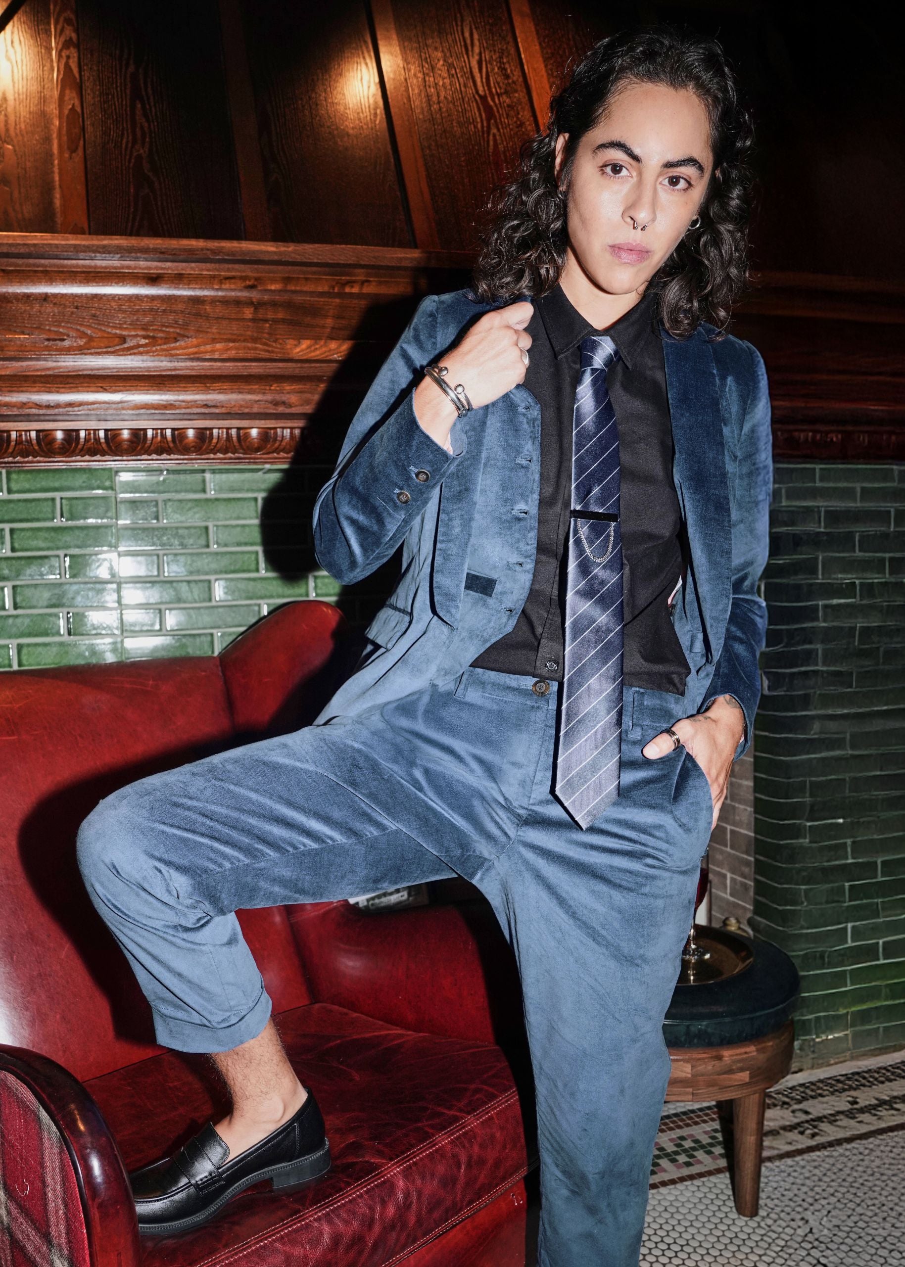 Person in blue velvet suit and tie looks at camera with one leg resting on red armchair.