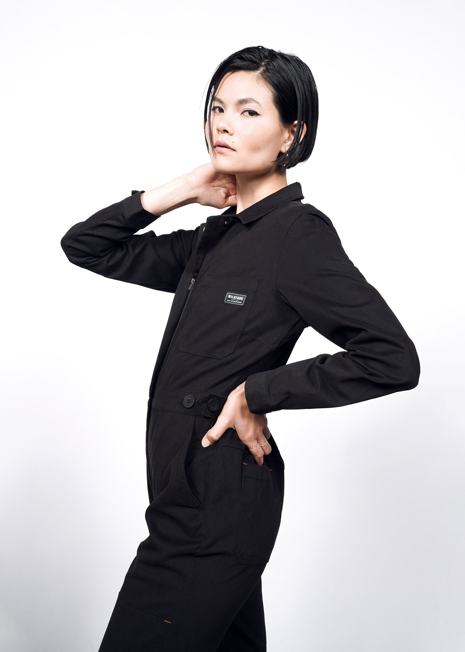 A person with short dark hair models The Essential Long Sleeve High Waisted Coverall in black, posing confidently with one hand on their hip and the other at their neck against a plain white background.