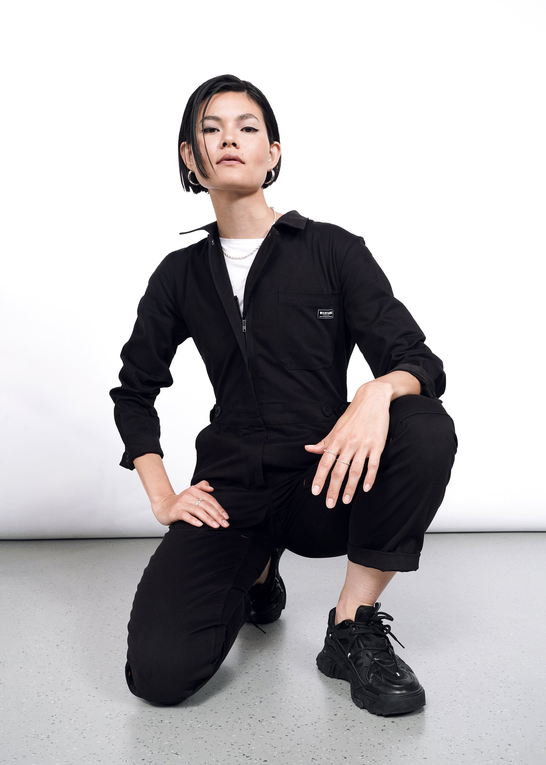 A person with short dark hair models The Essential Long Sleeve High Waisted Coverall with deep pockets and black sneakers, kneeling confidently on one knee against a plain white background.