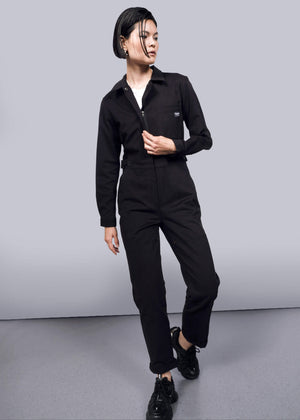 S=A person with short, wet-look hair models The Essential Long Sleeve High Waisted Coverall in black with deep pockets and black sneakers, standing against a plain white background, looking sideways with one hand on the zipper.