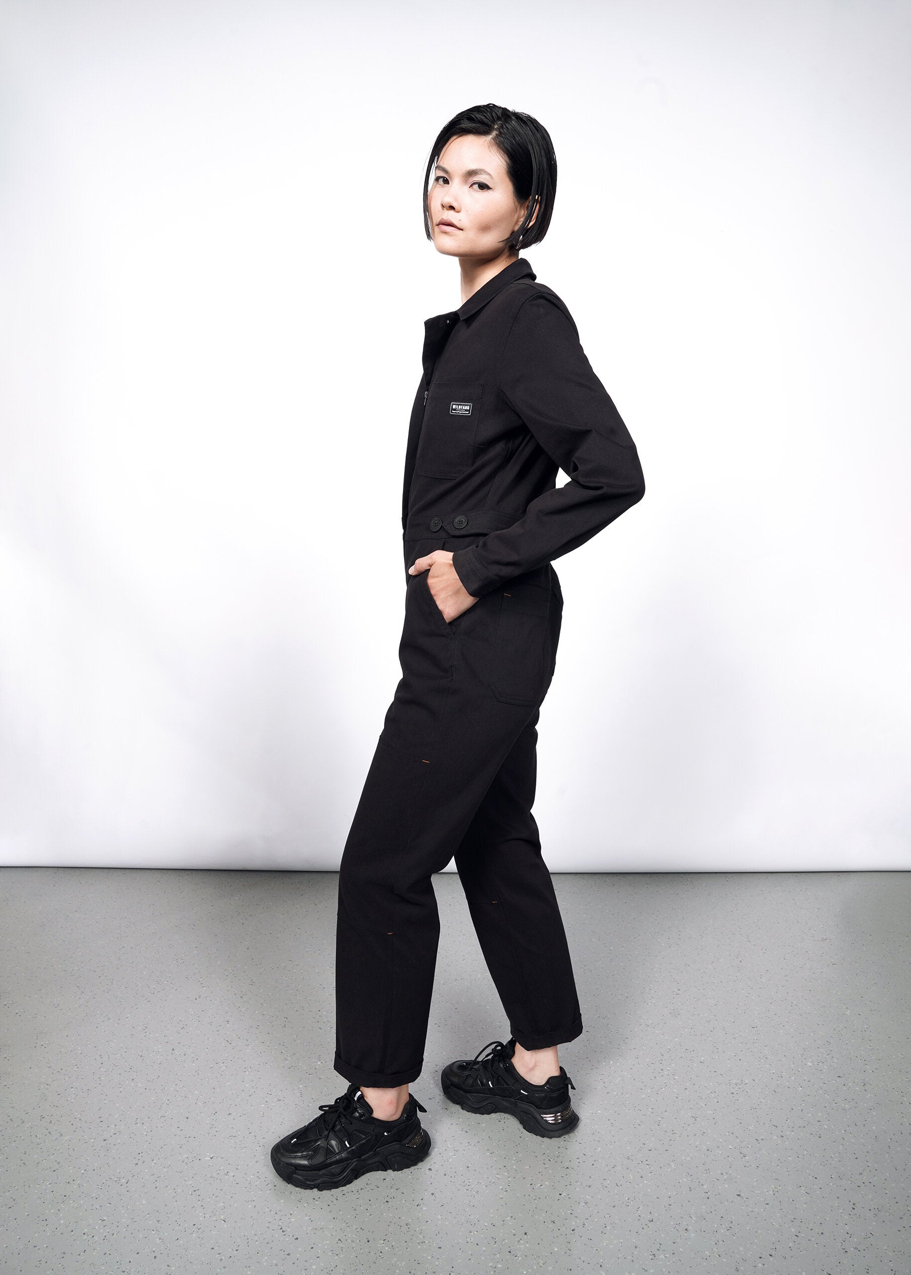 A person with short dark hair stands sideways, looking at the camera. They wear The Essential Long Sleeve High Waisted Coverall and black sneakers, posing against a plain white background and gray floor.