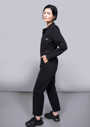 S=A person with short dark hair stands sideways, looking at the camera. They wear The Essential Long Sleeve High Waisted Coverall and black sneakers, posing against a plain white background and gray floor.