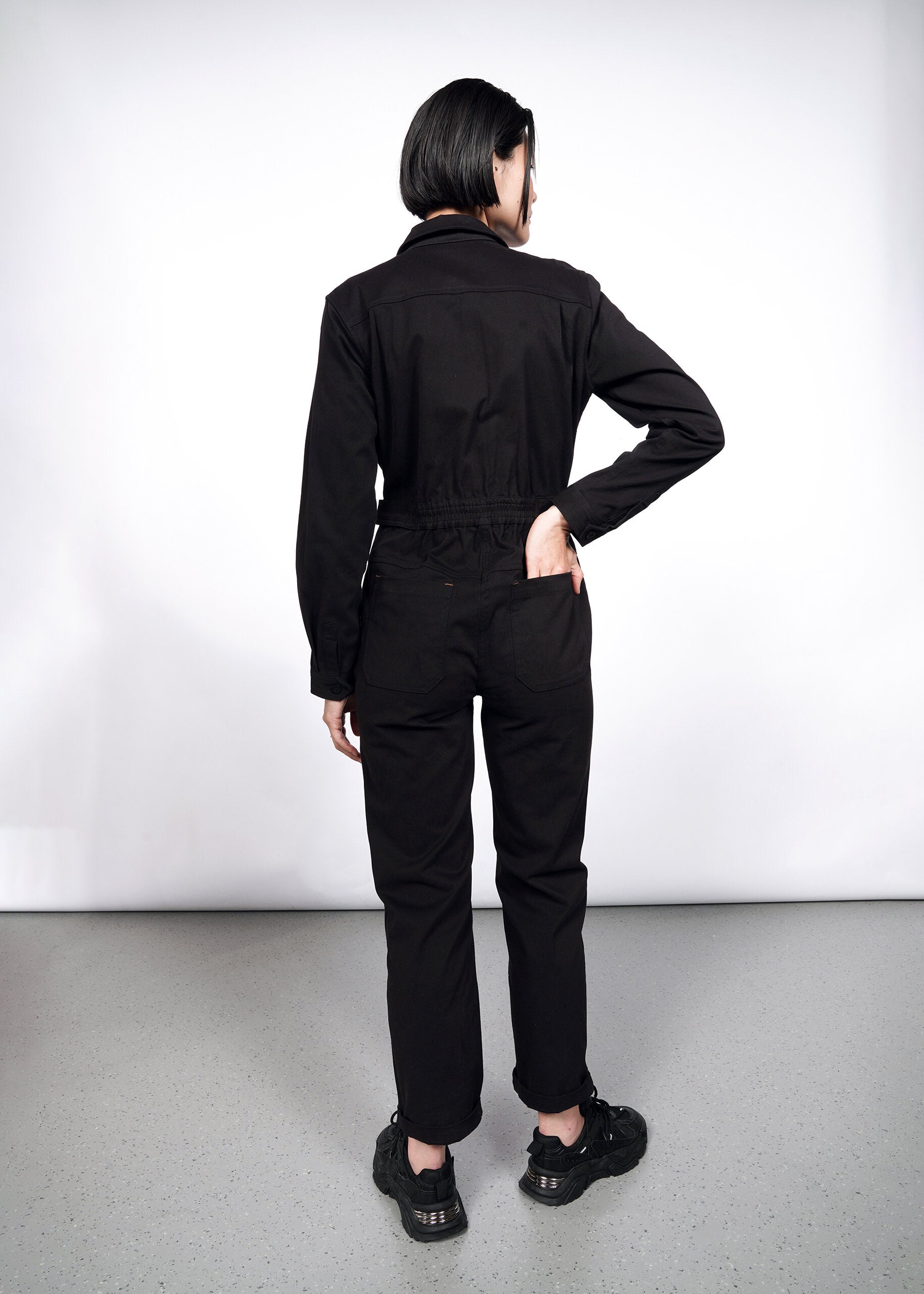 A person with short dark hair faces away, wearing The Essential Long Sleeve High Waisted Coverall and black sneakers, one hand on their hip and the other in a back pocket, set against a plain white background.