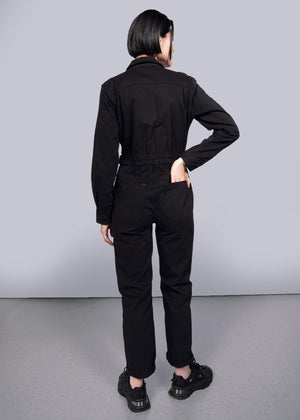 S=A person with short dark hair faces away, wearing The Essential Long Sleeve High Waisted Coverall and black sneakers, one hand on their hip and the other in a back pocket, set against a plain white background.