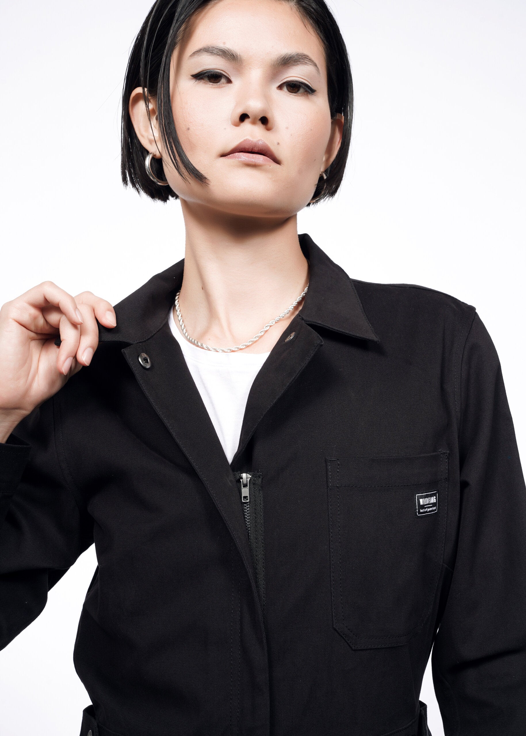 A person with short black hair wears The Essential Long Sleeve High Waisted Coverall in black over a white top, holding the collar. The background is plain white.