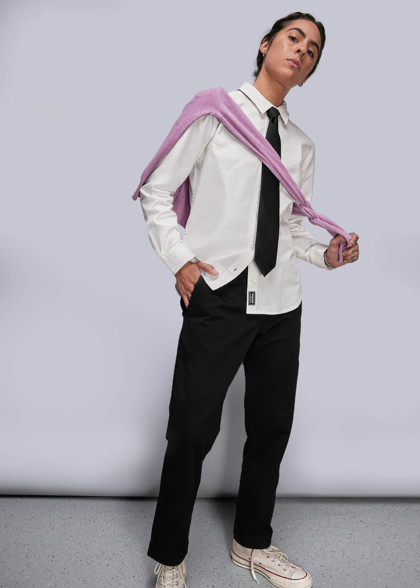 S=Person wearing a white shirt, black pants, and a pink sweater around shoulders with a gray background