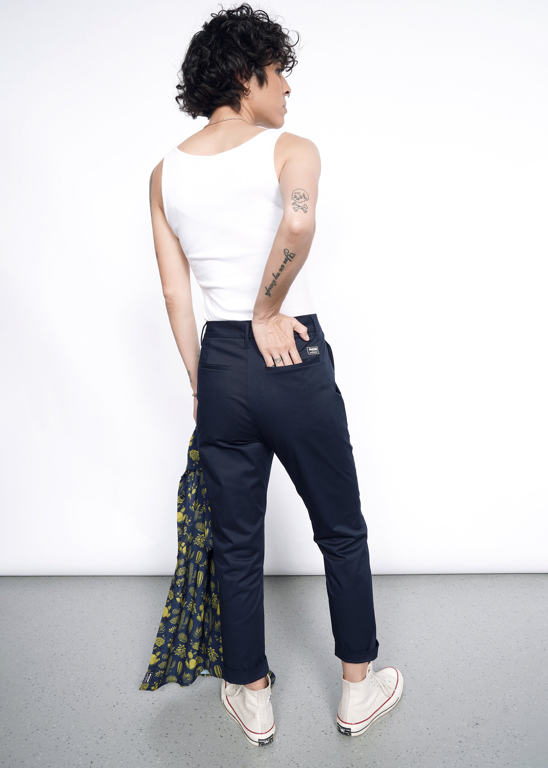 A person with short curly hair, wearing a white tank top, The Essential Slim Crop Pant in a tailored mid-rise fit, and white sneakers faces away holding a patterned jacket. Tattoos show on their left arm as they stand on a gray floor against a white background.