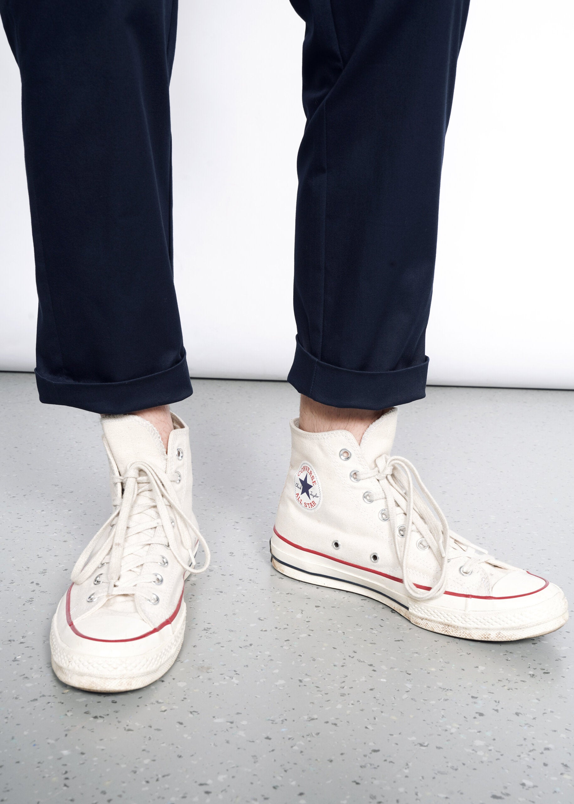 A person wears The Essential Slim Crop Pant in navy blue with a tailored mid-rise fit and off-white high-top Converse sneakers, standing on a speckled gray floor. One sneaker is loosely laced, with an untied lace trailing on the ground.