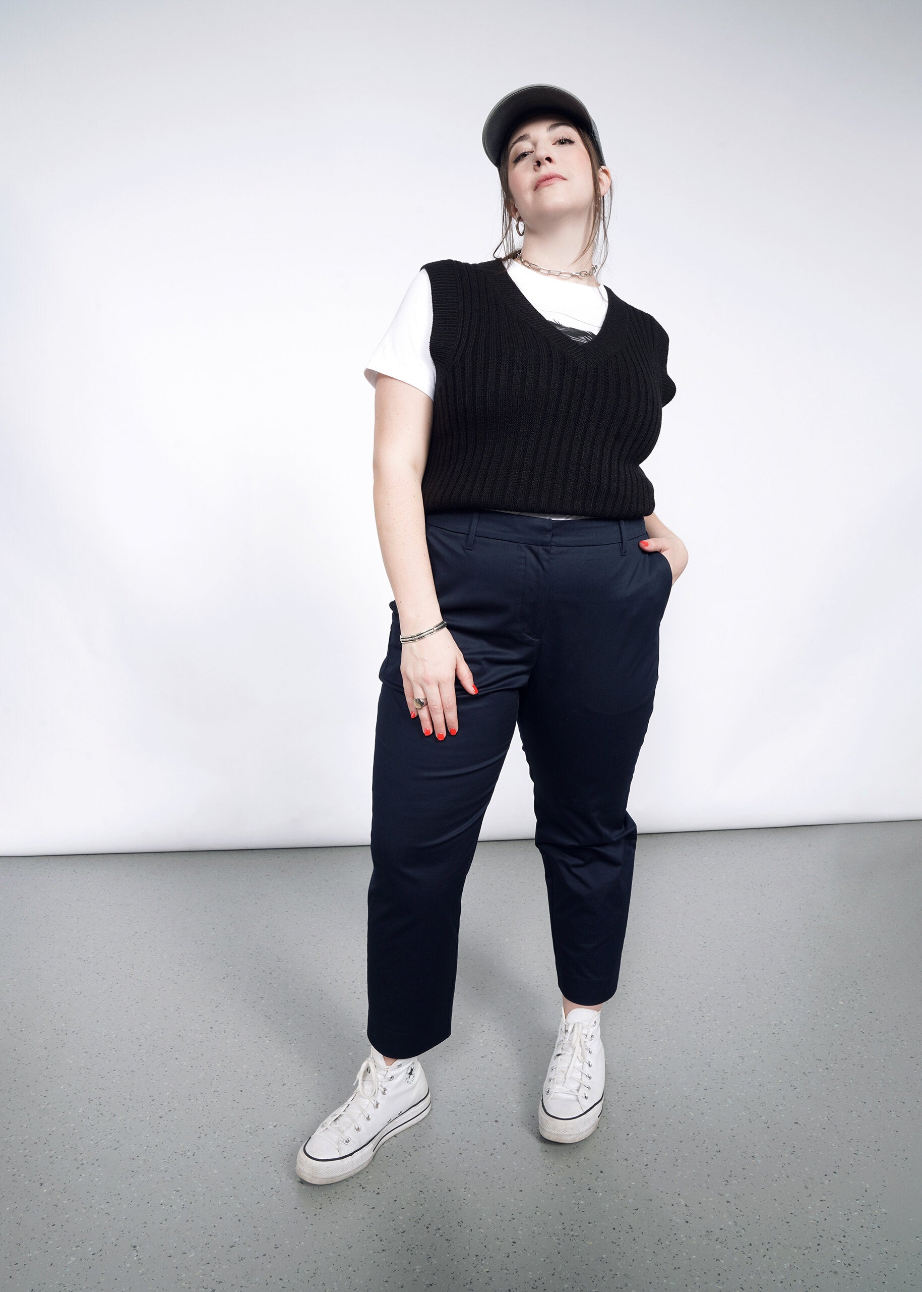 A person stands confidently against a plain background, wearing a gray cap, white T-shirt, black sweater vest, The Essential Slim Crop Pant with a tailored mid-rise fit, and white sneakers, one hand in their pocket and the other at their side.