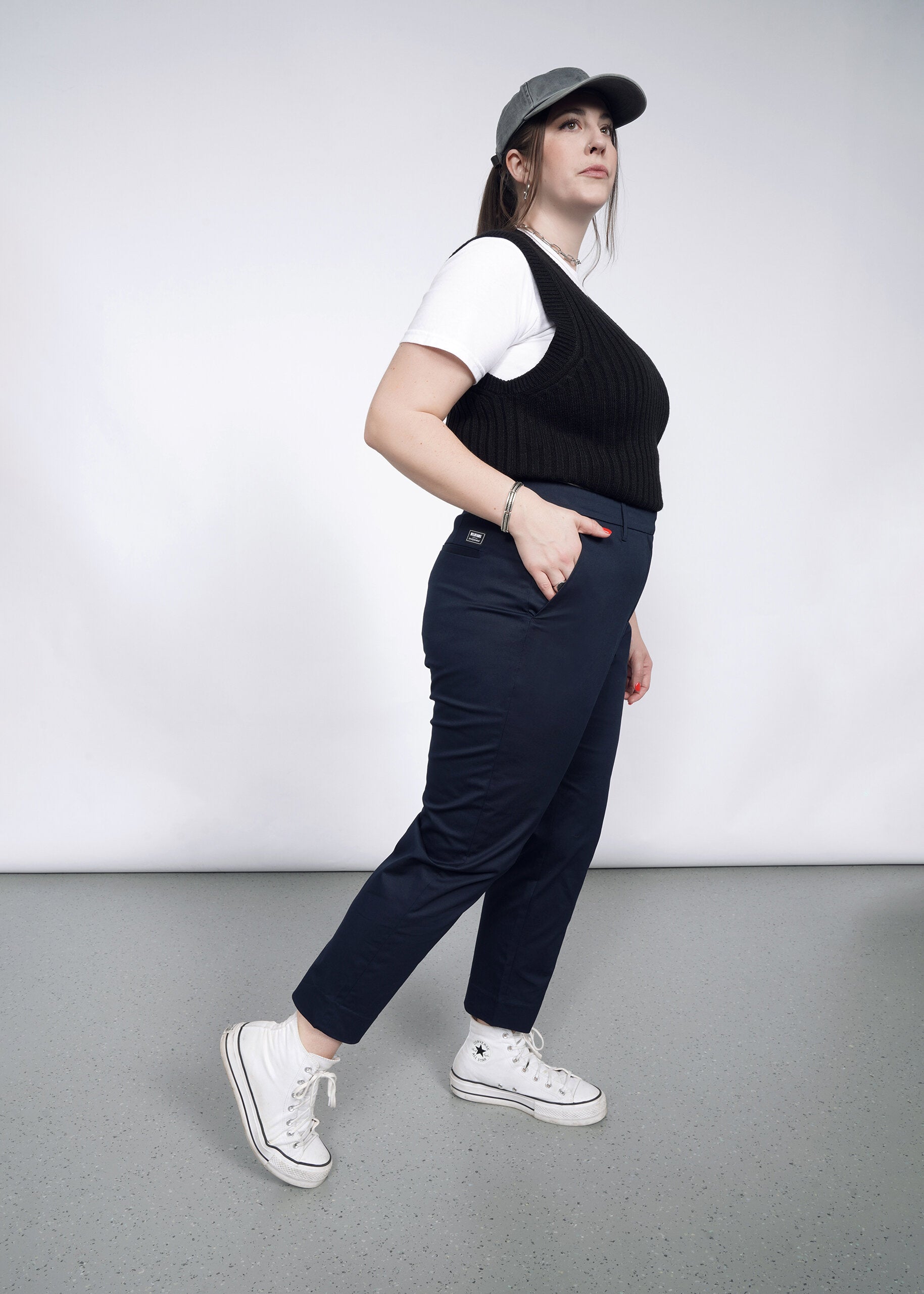 A person stands sideways against a plain background, wearing a gray cap, white t-shirt, black knit vest, and The Essential Slim Crop Pant in navy for a tailored mid-rise fit. White sneakers complete the look as they gaze slightly upward with one hand in their pocket.