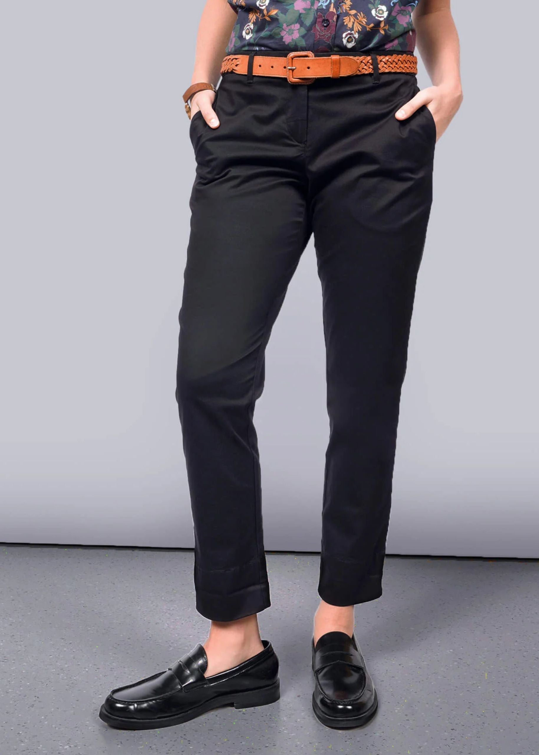 Pictured from waist down, person wears black trouser with brown belt and black loafers on grey background.