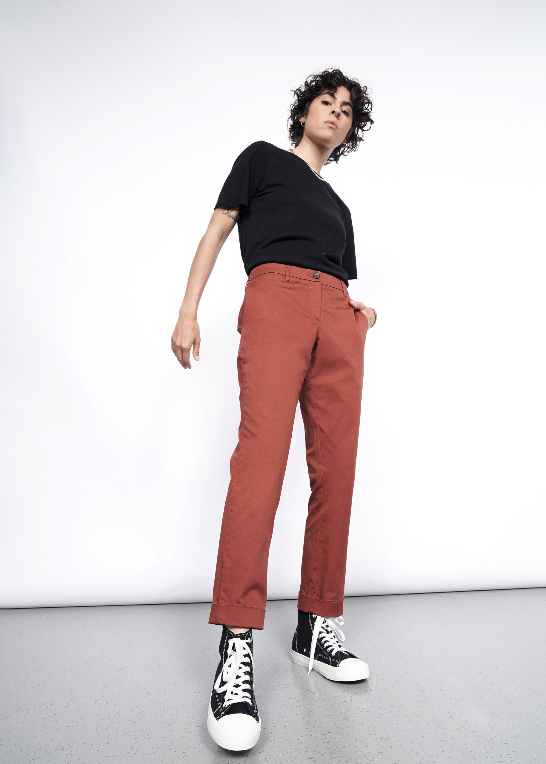A person with short curly hair poses confidently in a black t-shirt, The Essential Trouser in rust (relaxed fit), and black high-top sneakers against a white background. The low-angle shot highlights their strong stance and classic style.