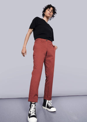 S=A person with short curly hair poses confidently in a black t-shirt, The Essential Trouser in rust (relaxed fit), and black high-top sneakers against a white background. The low-angle shot highlights their strong stance and classic style.