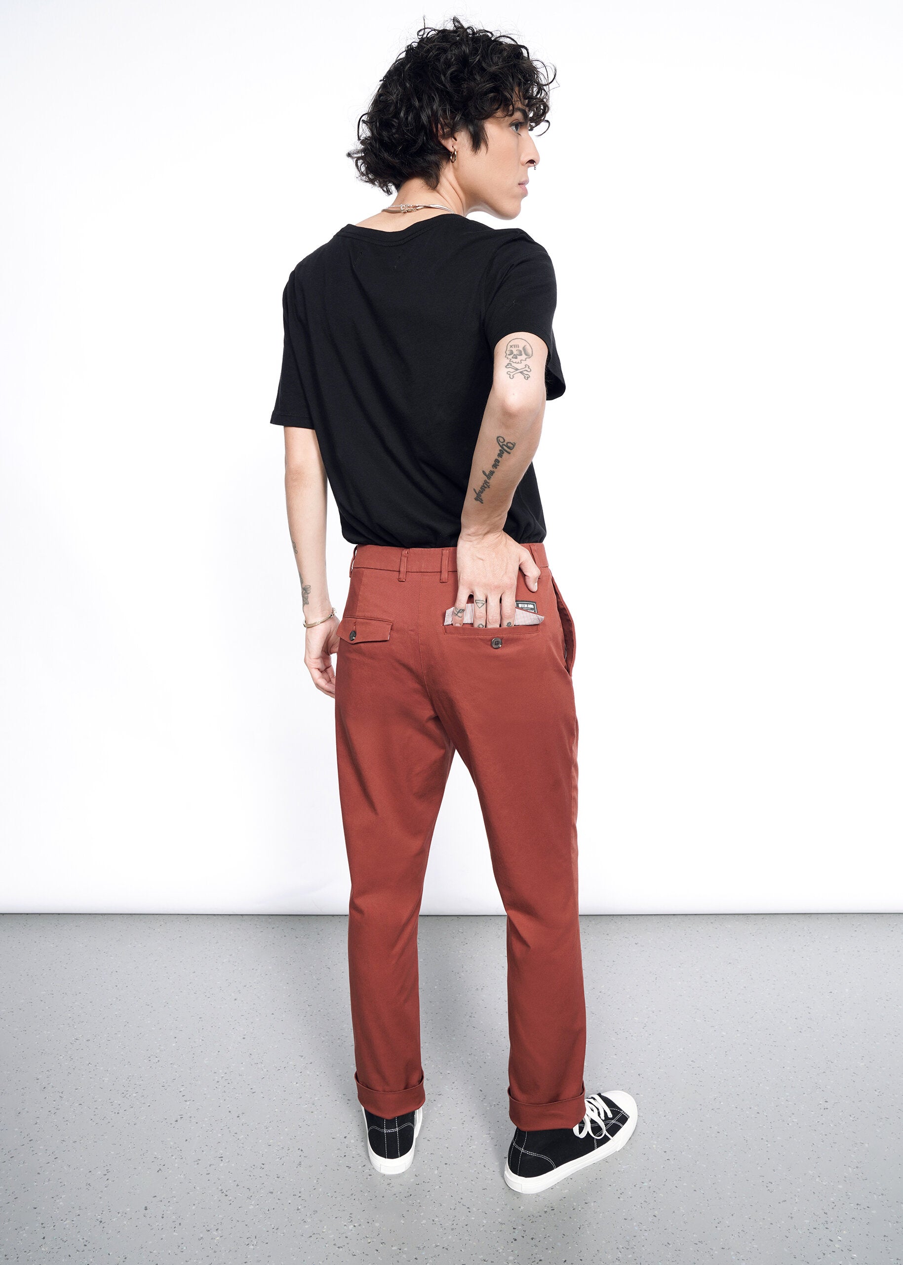 Wearing The Essential Trouser in a relaxed rust color, a person with short curly hair, black T-shirt, and black sneakers stands turned away with their right hand in the back pocket and tattoos showing on their left arm.