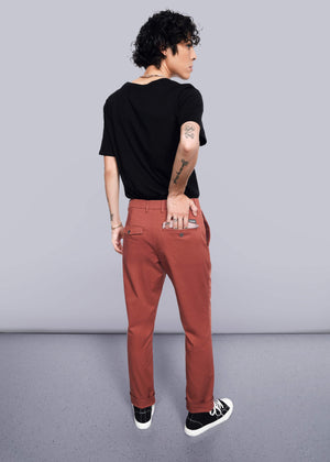 S=Wearing The Essential Trouser in a relaxed rust color, a person with short curly hair, black T-shirt, and black sneakers stands turned away with their right hand in the back pocket and tattoos showing on their left arm.