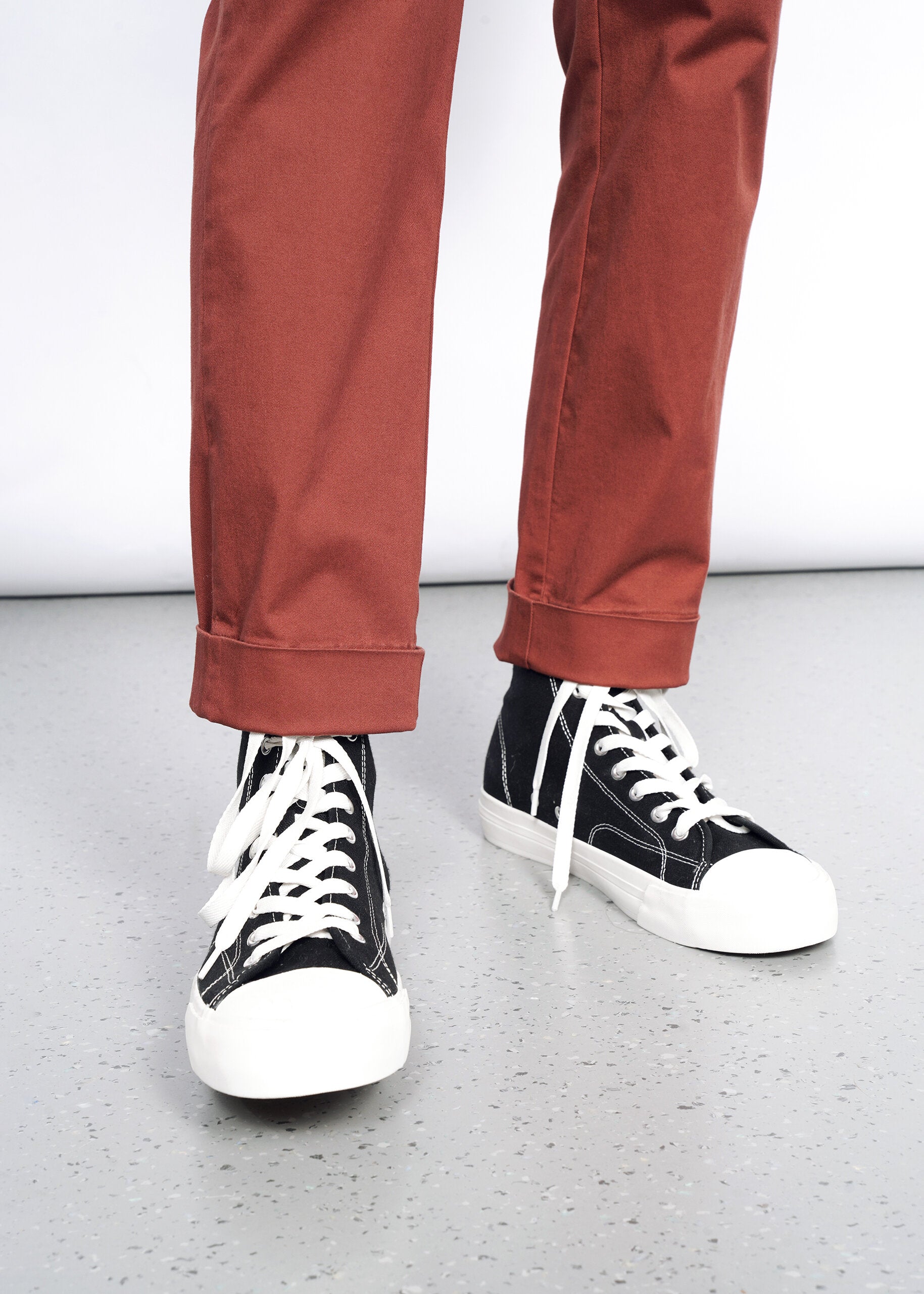 Wearing The Essential Trouser in a relaxed rust hue—timeless wardrobe staples—paired with black high-top sneakers featuring white soles and laces, standing on a light gray speckled floor against a plain background.