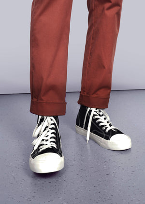 S=Wearing The Essential Trouser in a relaxed rust hue—timeless wardrobe staples—paired with black high-top sneakers featuring white soles and laces, standing on a light gray speckled floor against a plain background.