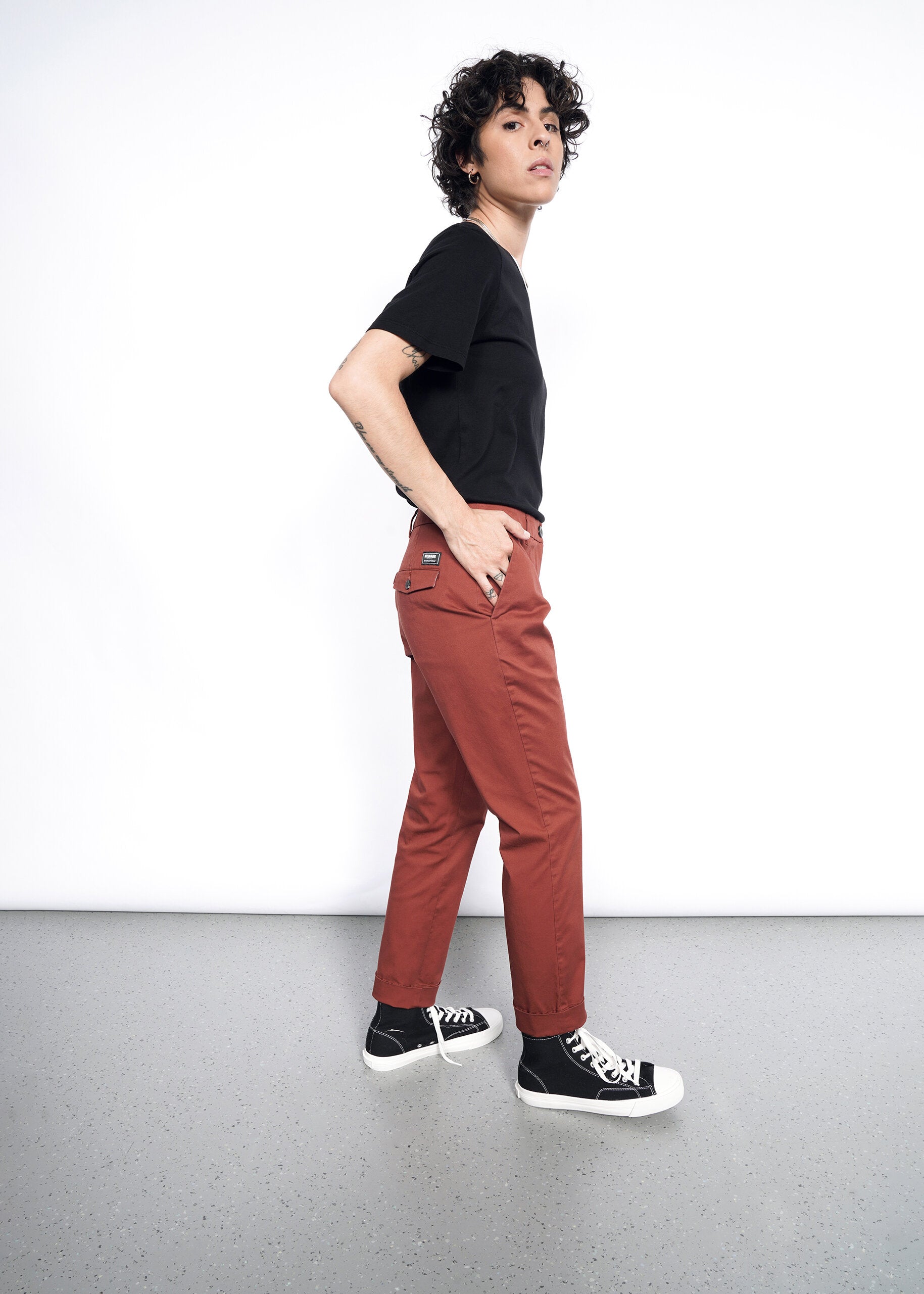 Person with short curly hair stands sideways against a white wall, wearing a black t-shirt, The Essential Trouser in rust, and black high-top sneakers with white toes, looking confidently at the camera—a true wardrobe staple.
