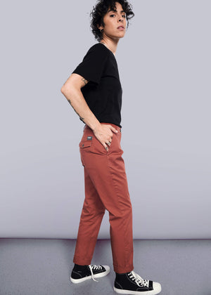 S=Person with short curly hair stands sideways against a white wall, wearing a black t-shirt, The Essential Trouser in rust, and black high-top sneakers with white toes, looking confidently at the camera—a true wardrobe staple.