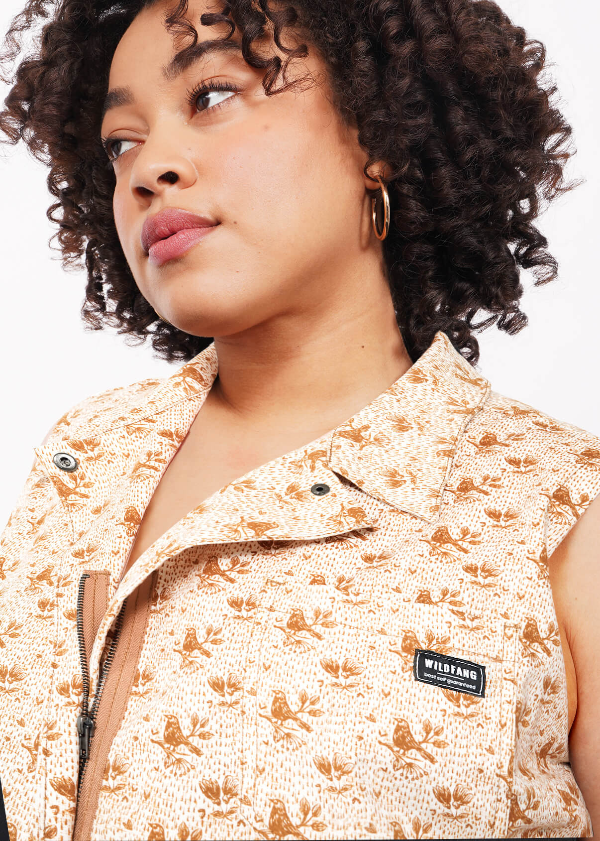 A person with curly hair wears The Essential Sleeveless High Waisted Coverall in beige with an adjustable waist and black Wildfang label, paired with gold hoop earrings, looking to the side against a plain white background.