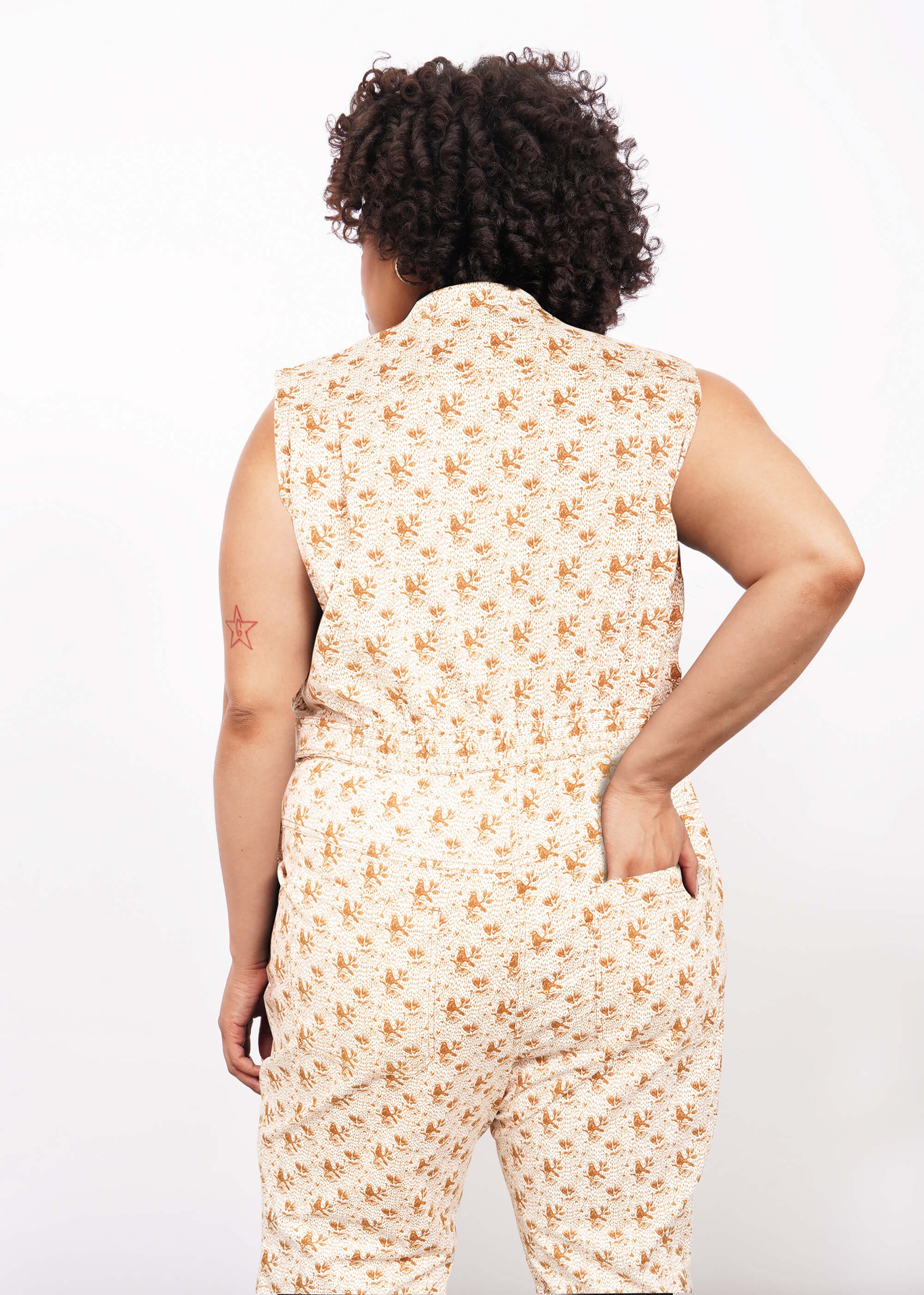 A person with curly hair, seen from behind, wears The Essential Sleeveless High Waisted Coverall in cream with a floral design. Their left hand rests in a back pocket, revealing a red star tattoo on their upper arm. Background is white.