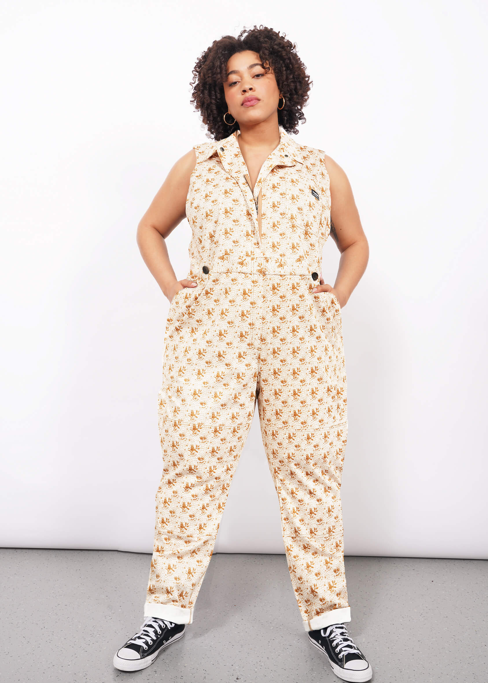 A woman with curly hair models The Essential Sleeveless High Waisted Coverall, showcasing a tan and brown floral pattern, collared design, deep pockets, and high-waisted fit. She pairs it with black and white sneakers against a white background.