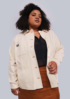 1X=A person with curly hair poses confidently, wearing The Essential Denim Utility Jacket over a black top and a brown skirt. The cream-colored jacket features deep pockets and a label, set against a plain, light background.