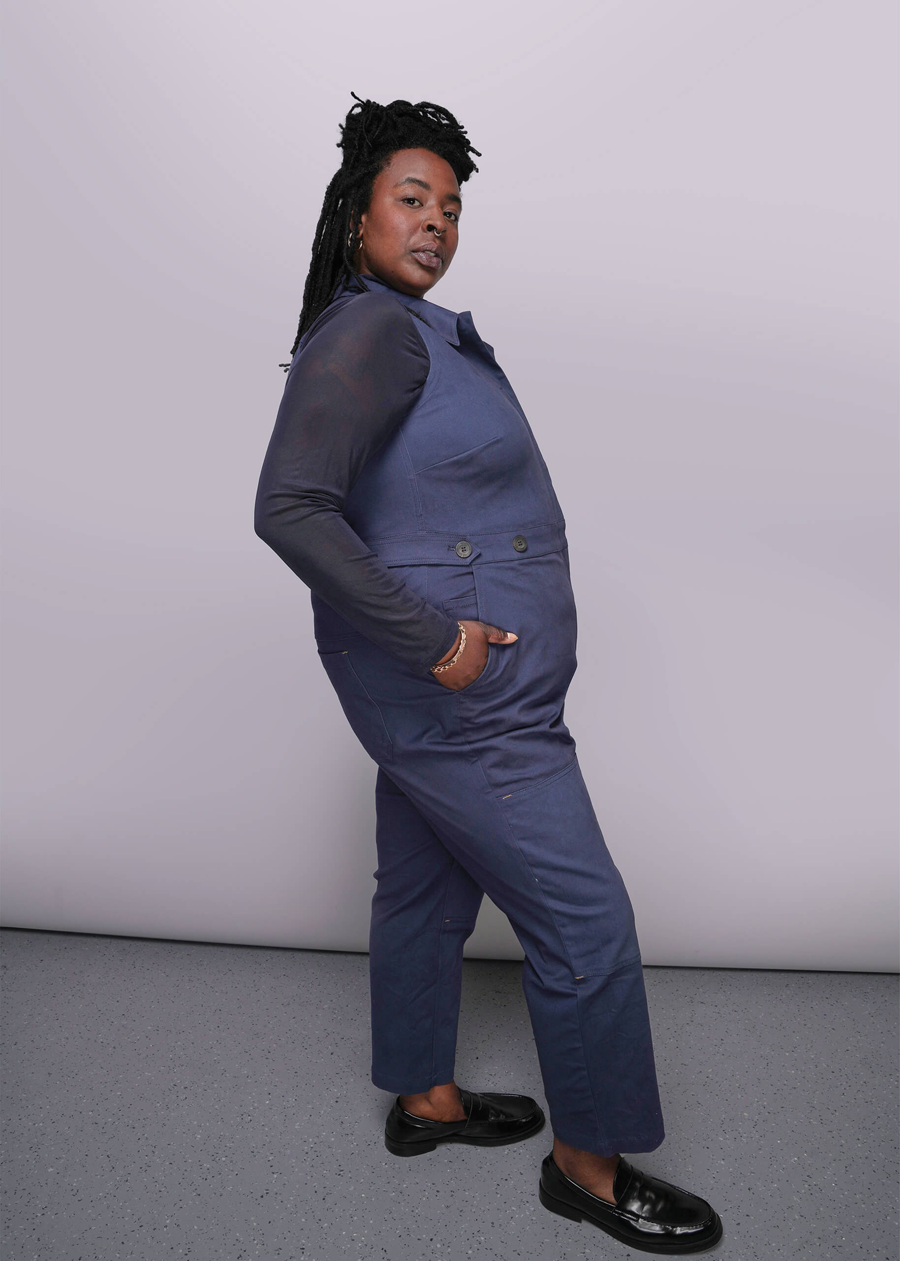1X=Person wearing a blue coverall with hand in side pocket against a plain background
