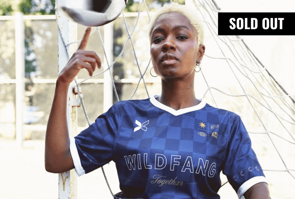 Model with short curly blonde hair spins soccer ball on their finger while looking at camera. They are wearing the Togethxr x Wildfang Jersey. A banner in the top right corner reads SOLD OUT.