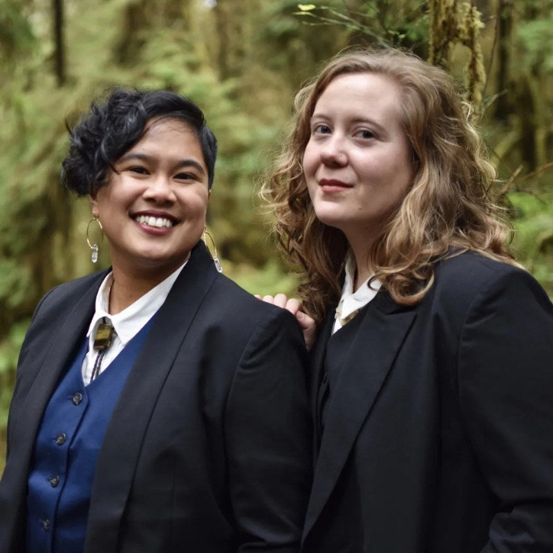 Two people wearing black suits pose in from of natural backdrop smiling at camera.
