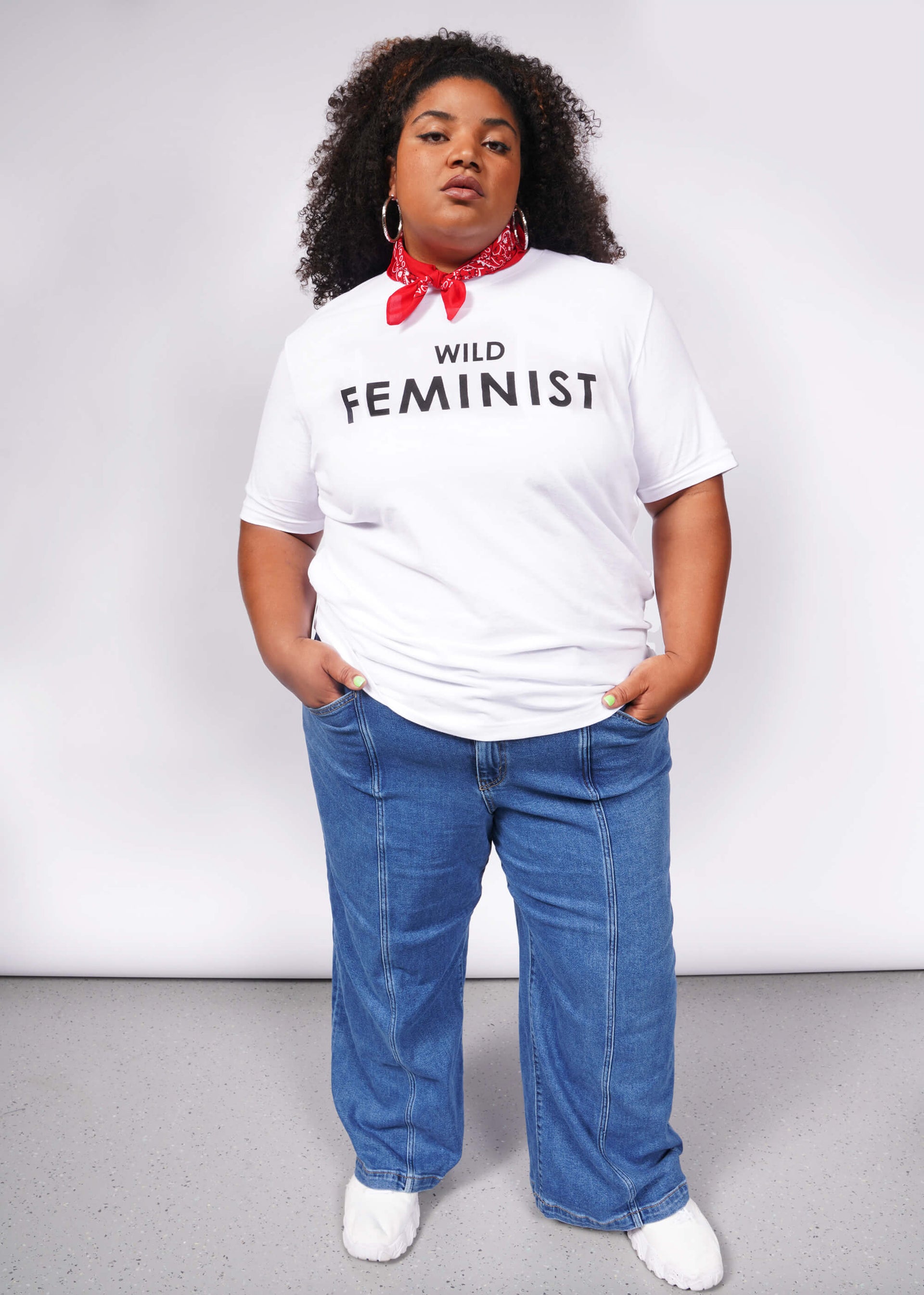 A confident person wears The Wild Feminist® Tee, a relaxed unisex fit white shirt, paired with a red bandana, blue jeans, and white shoes. The plain background emphasizes their commitment to unity and advocacy.