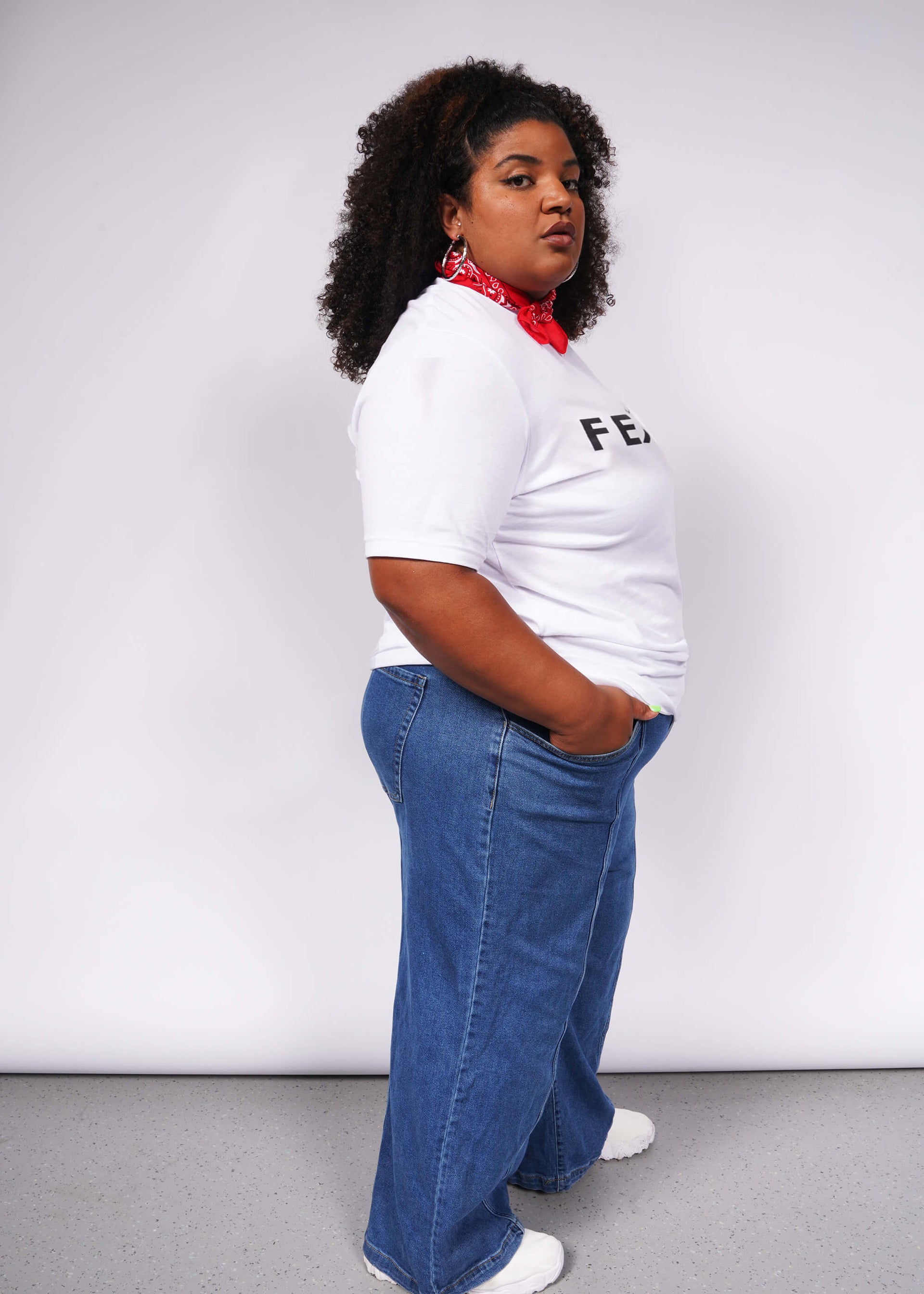 A person with curly hair confidently poses against a gray background, wearing a relaxed unisex fit. They sport The Wild Feminist® Tee, blue jeans, white sneakers, and a red bandana around their neck. One hand in their pocket, they embody unity.