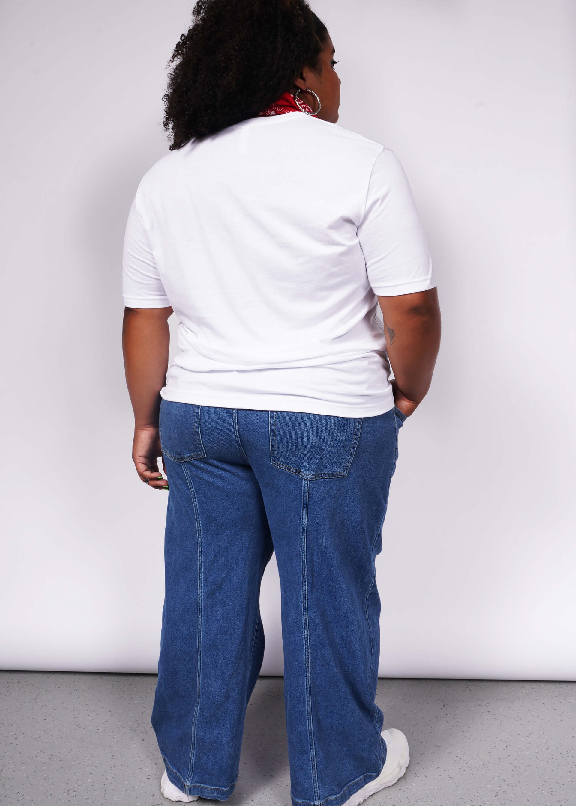 A person with curly hair and relaxed poise stands with their back to the camera in a unisex white Wild Feminist® Tee and blue jeans, accented by a red scarf. The plain light gray background highlights their effortless style.