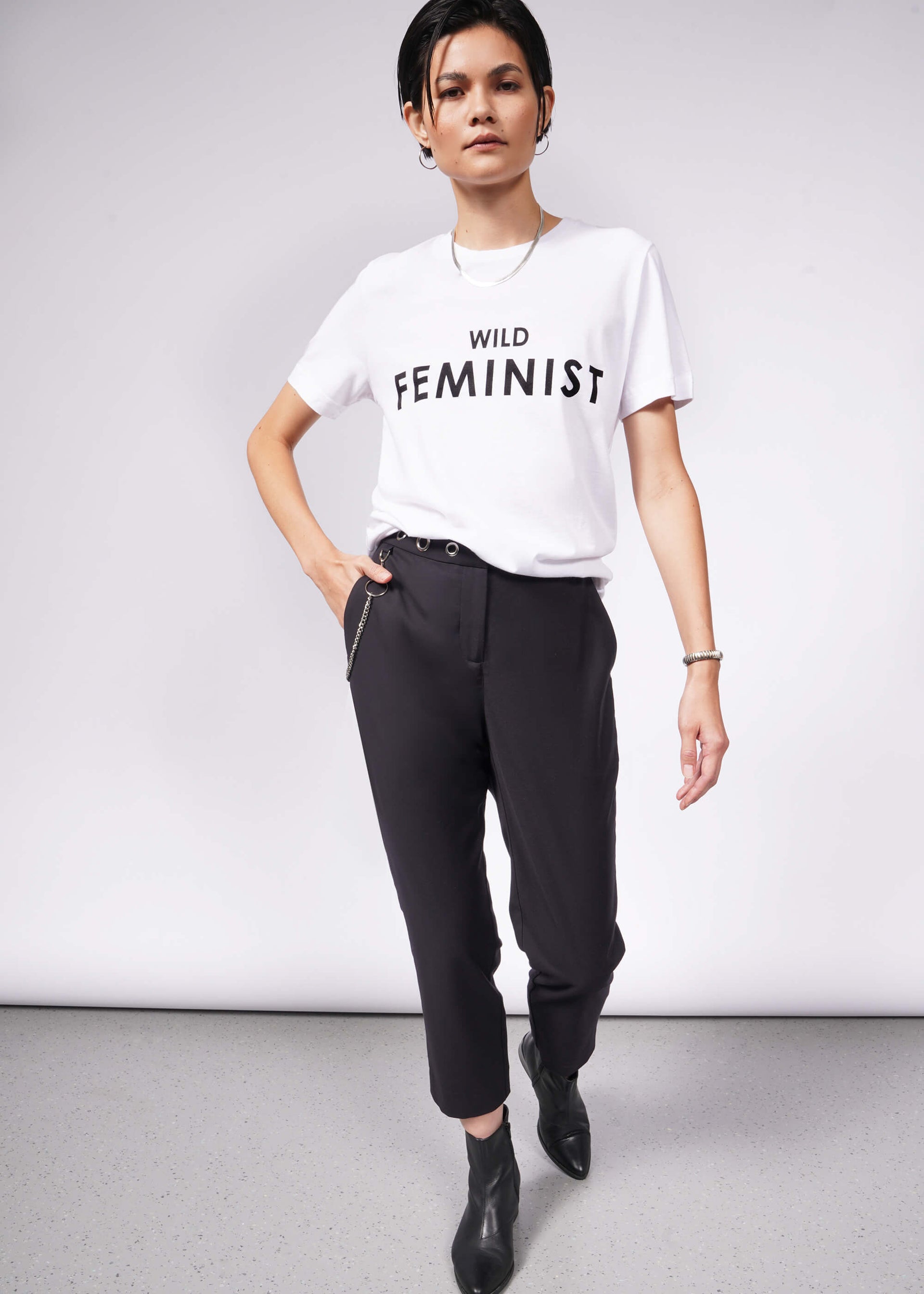 A person with short dark hair wears The Wild Feminist® Tee, black pants, and black ankle boots, embodying a relaxed unisex fit. Standing confidently against a plain background with one hand in their pocket, they inspire unity.