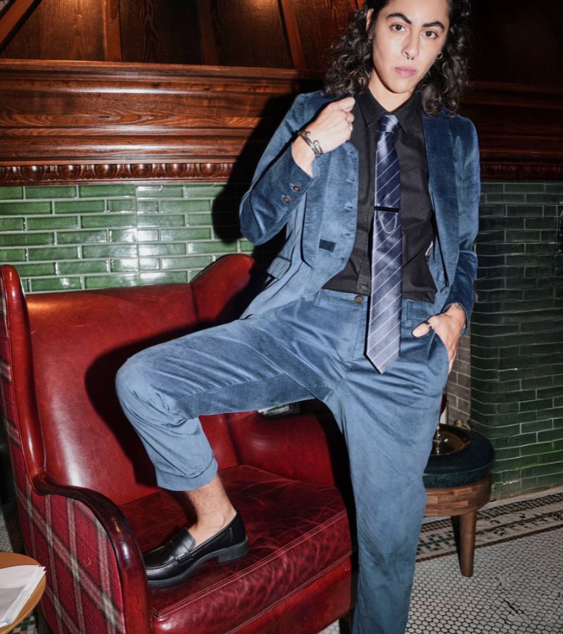Person wearing blue velvet suit looks at camera with leg resting on red leather armchair.
