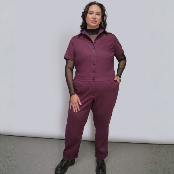 Person standing against grey background wearing burgundy colored jumpsuit with black mesh top underneath and black boots.