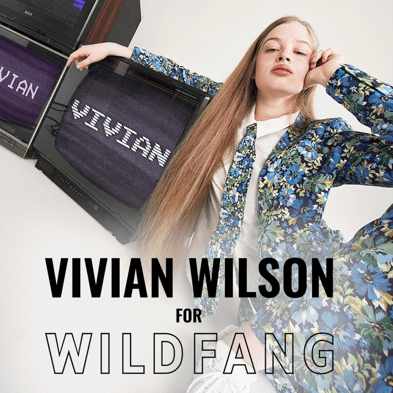 Vivian Wilson sits on floor resting her arm on a vintage TV displaying her name, vivian, on them. She's wearing The Empower Tux Blazer, The Empower Tie, and The Empower Wide Leg Pant all in Carnations Navy, a blue toned floral print. Text overlay on image reads VIVIAN WILSON FOR WILDFANG.