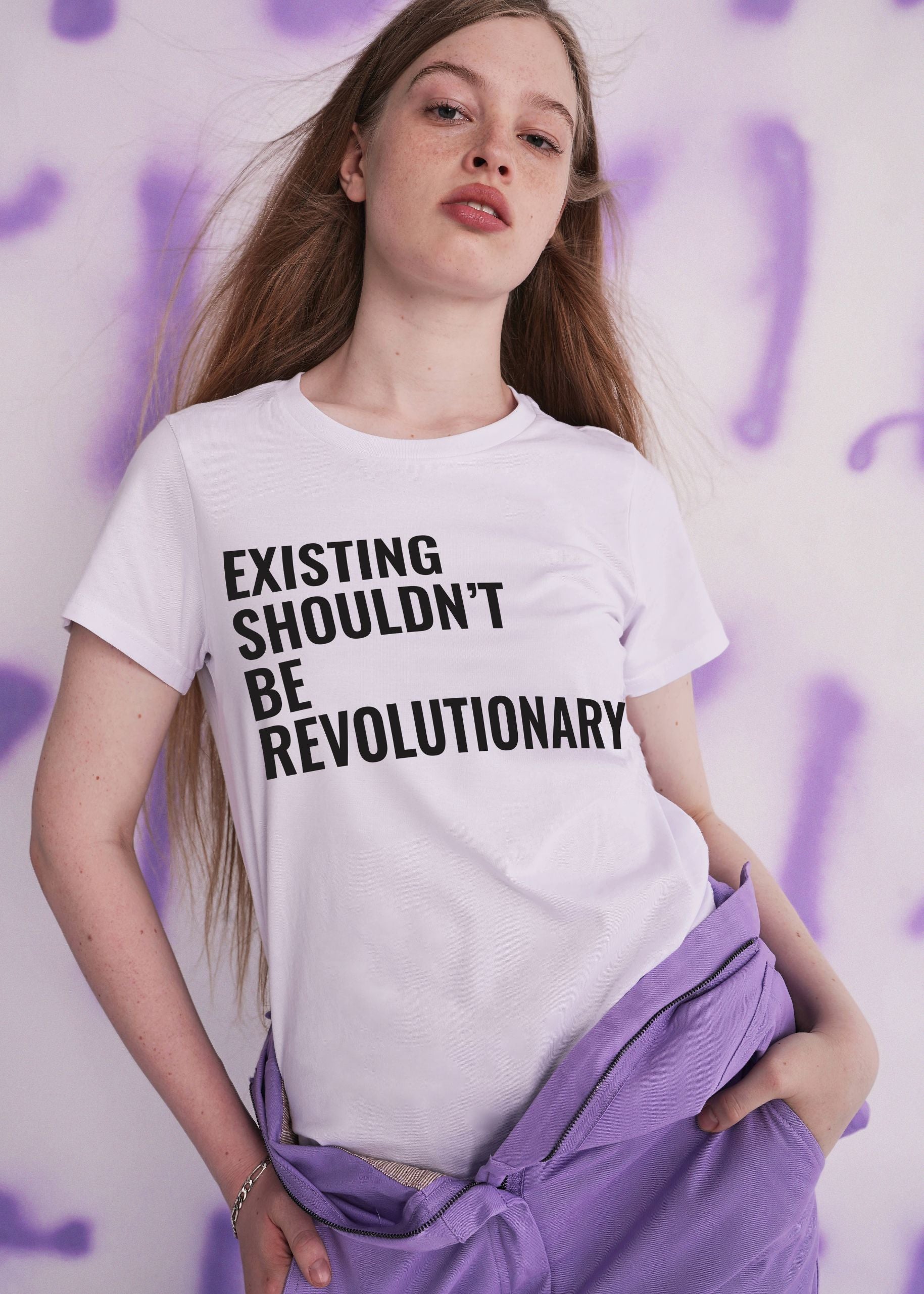 Vivian Wilson models the VIVIAN WILSON FOR WILDFANG TEE, featuring bold EXISTING SHOULDNT BE REVOLUTIONARY text, paired with purple coveralls worn half on against a white wall marked by purple spray paint.