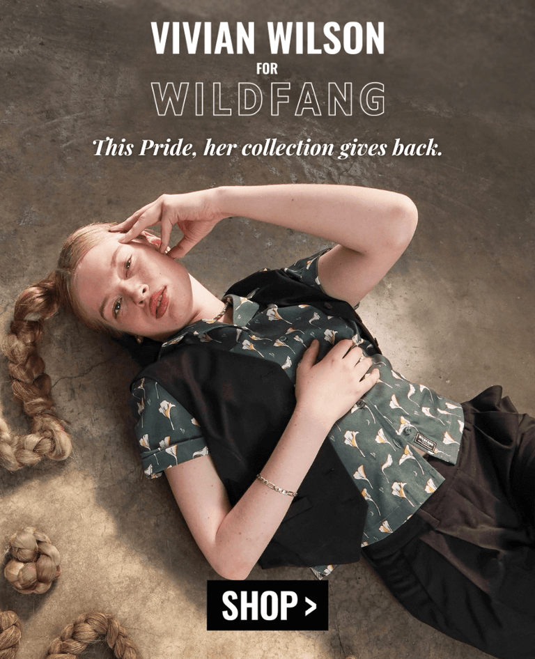 Vivian Wilson sits on floor of room with white walls and concrete floor, she wears The Empower Vest, The Everyday Camp Button Up, and The Empower Short. Her hair is braided. Text overlay reads "VIVIAN WILSON FOR WILDFANG. This pride her collection gives back to The Trevor Project." A button below reads SHOP
