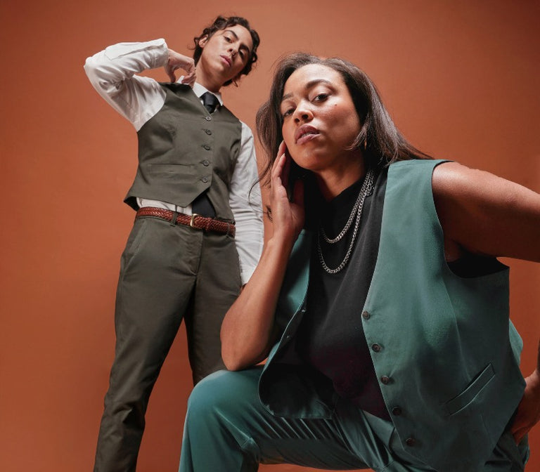 Two models wearing matching vest and pant suits look at camera while standing in front of orange gradient backdrop.